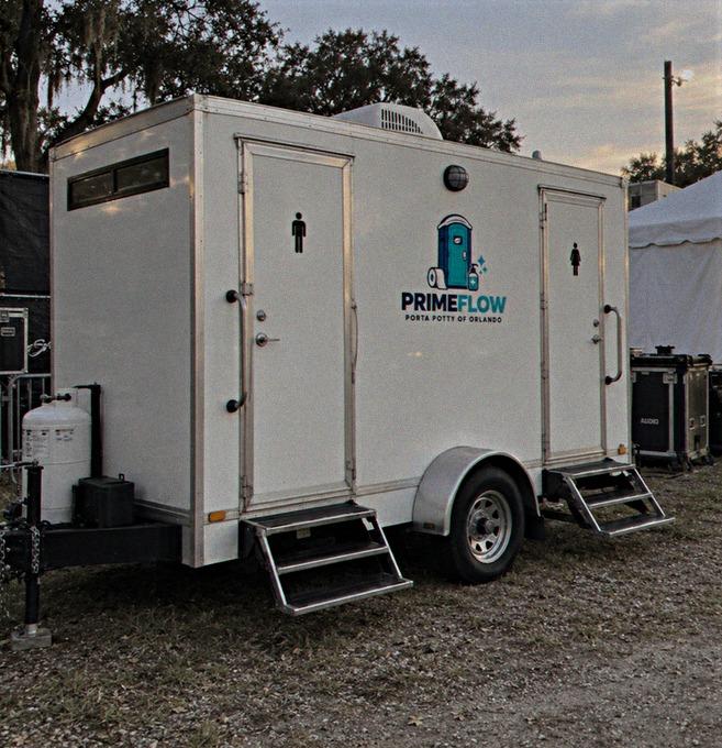 White two-door restroom trailer with fold-out steps parked beside event tents