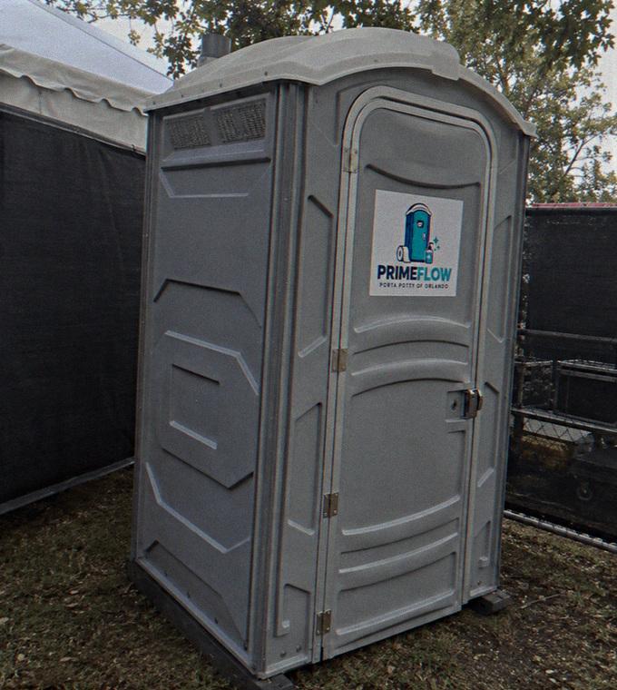 Grey portable restroom standing on grass beside a site fence